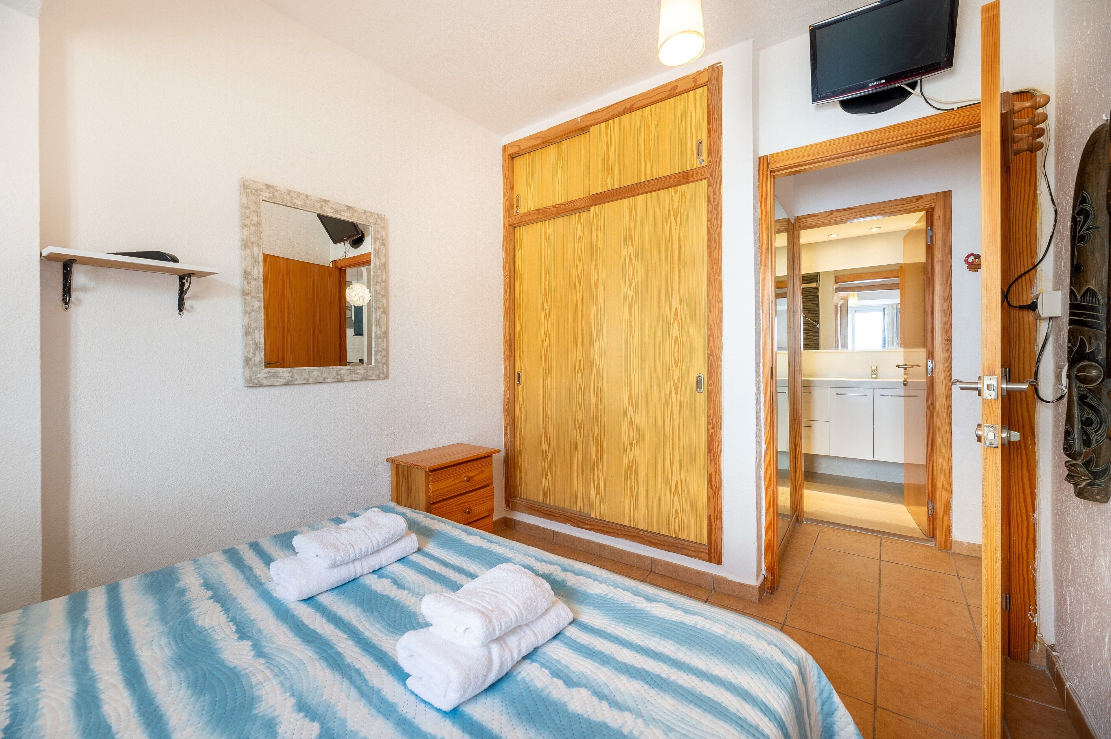 Une chambre à coucher avec un lit, une armoire en bois, un téléviseur et une salle de bain visible à travers une porte ouverte.