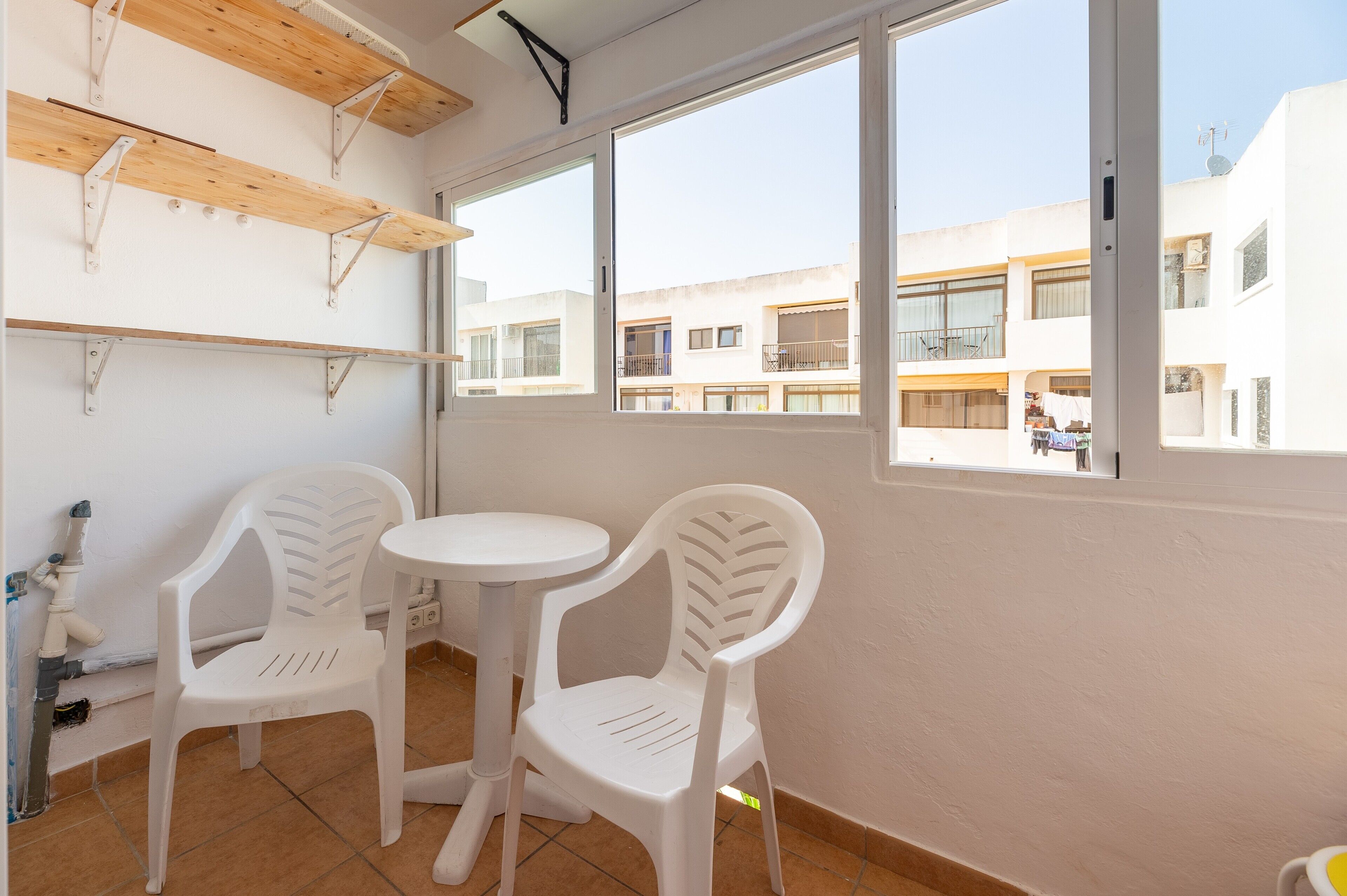 Un balcón con dos sillas de plástico blanco, una mesita redonda y vistas a edificios cercanos.