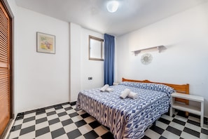 A bedroom with a checkered floor, a bed with patterned bedding, a window with blue curtains, a wall-mounted shelf, and a framed picture.