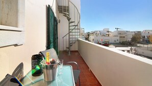 A balcony with a table, a bottle, and a glass, overlooking a street with buildings and a spiral staircase.
