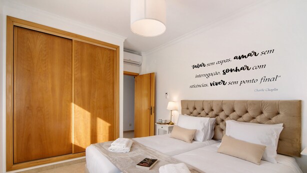 A hotel room with a large wooden wardrobe, a bed with white linens, a bedside table with a lamp, and a motivational quote on the wall.