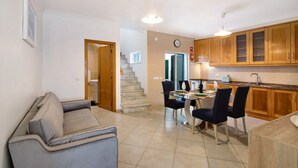 A modern kitchen with wooden cabinets, a dining area with a table and chairs, a grey sofa, and a staircase leading to another level.