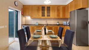 A modern kitchen with wooden cabinets, a stainless steel refrigerator, and a glass dining table with four chairs.
