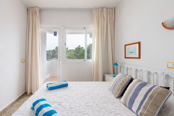 A bedroom with a bed, curtains, a window, a bedside table, and a wall-mounted light fixture.