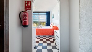 Habitación de hotel con piso a cuadros, cama con ropa de cama roja, cómoda blanca, extintor de incendios y balcón con vistas.