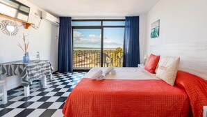 Habitación de hotel con piso a cuadros, cama con ropa de cama roja, escritorio con mantel a rayas y balcón con vistas.