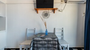 Una habitación con una televisión montada en un estante de madera, una mesa con mantel estampado, dos sillas blancas y un espejo decorativo en la pared.