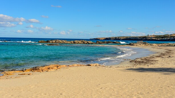 Una playa con aguas azul cristalinas, orilla arenosa y afloramientos rocosos.