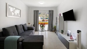 Ein modernes Wohnzimmer mit einer Couch, einem Flachbildfernseher, einem kleinen Tisch mit einer Pflanze und Blick auf einen Balkon mit Gartenmöbeln.