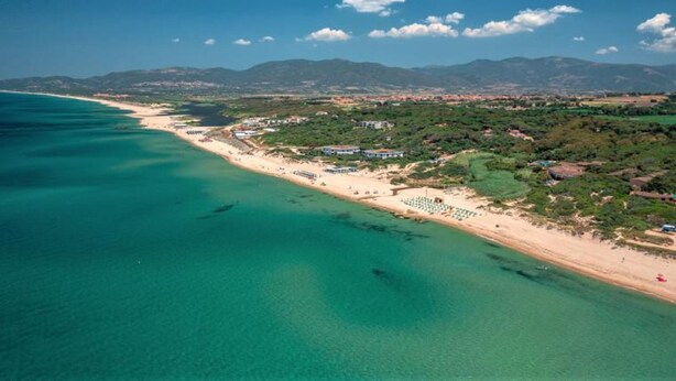 Vue aérienne d’une zone côtière avec une plage, des bâtiments et des montagnes en arrière-plan.