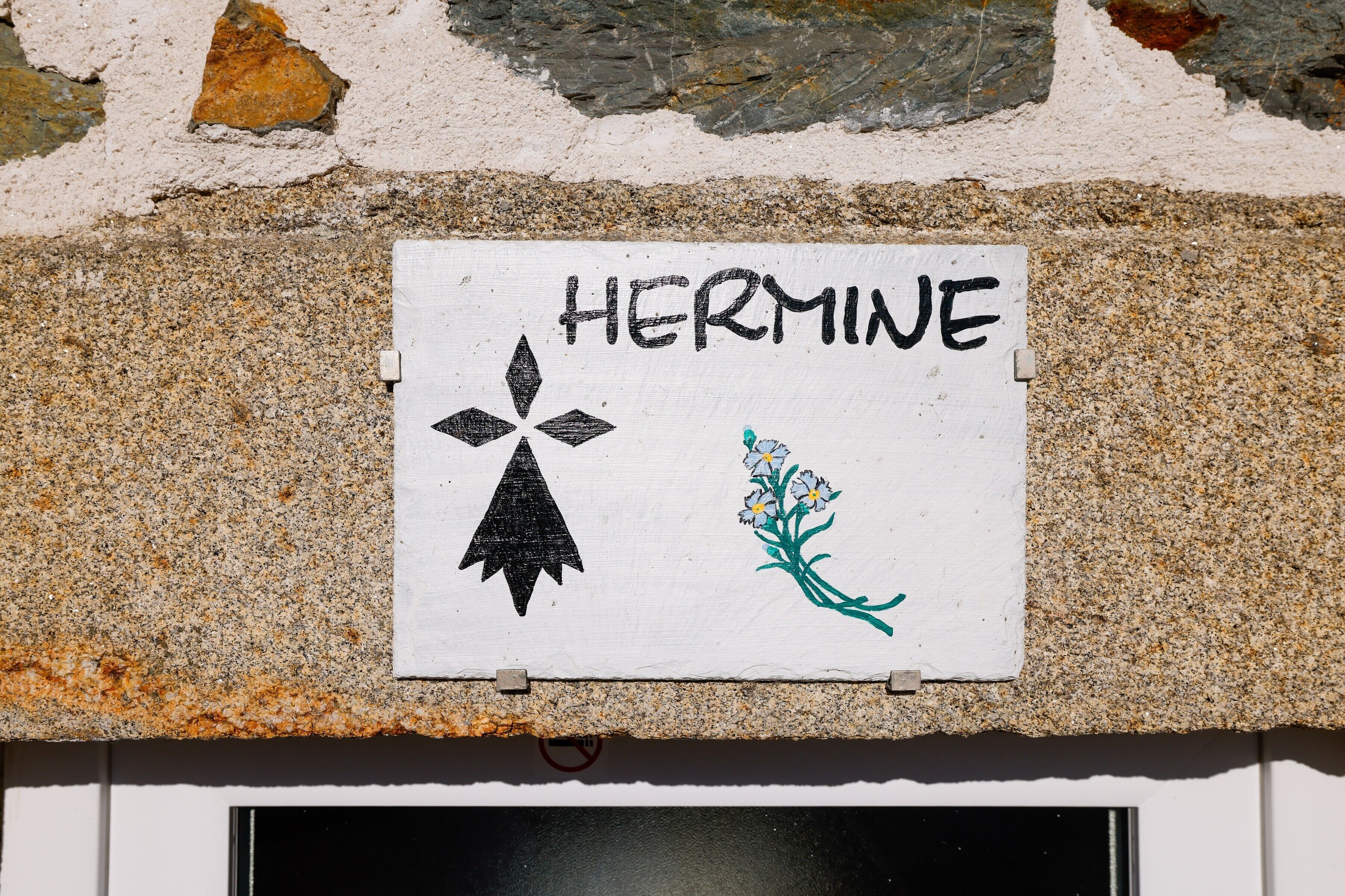 Ein Schild mit dem Wort 'HERMINE' und einem Symbol, an der Gebäudefassade angebracht.