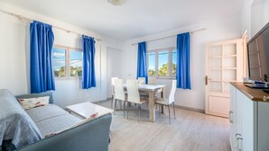 A modern living room with a sofa, dining table, and chairs, a kitchen area, and large windows with curtains. - Apartment 'Apartamento Los Hermanos' with Wi-Fi and Air Conditioning (Es Pujols)