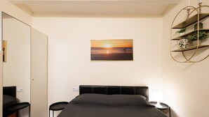 Ein Schlafzimmer mit Bett, Nachttischen, einem Wandregal mit Pflanzen und einem Sonnenuntergangsbild.