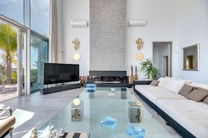 A modern living room with a large flat-screen TV, a glass coffee table, a white sofa, and a fireplace.