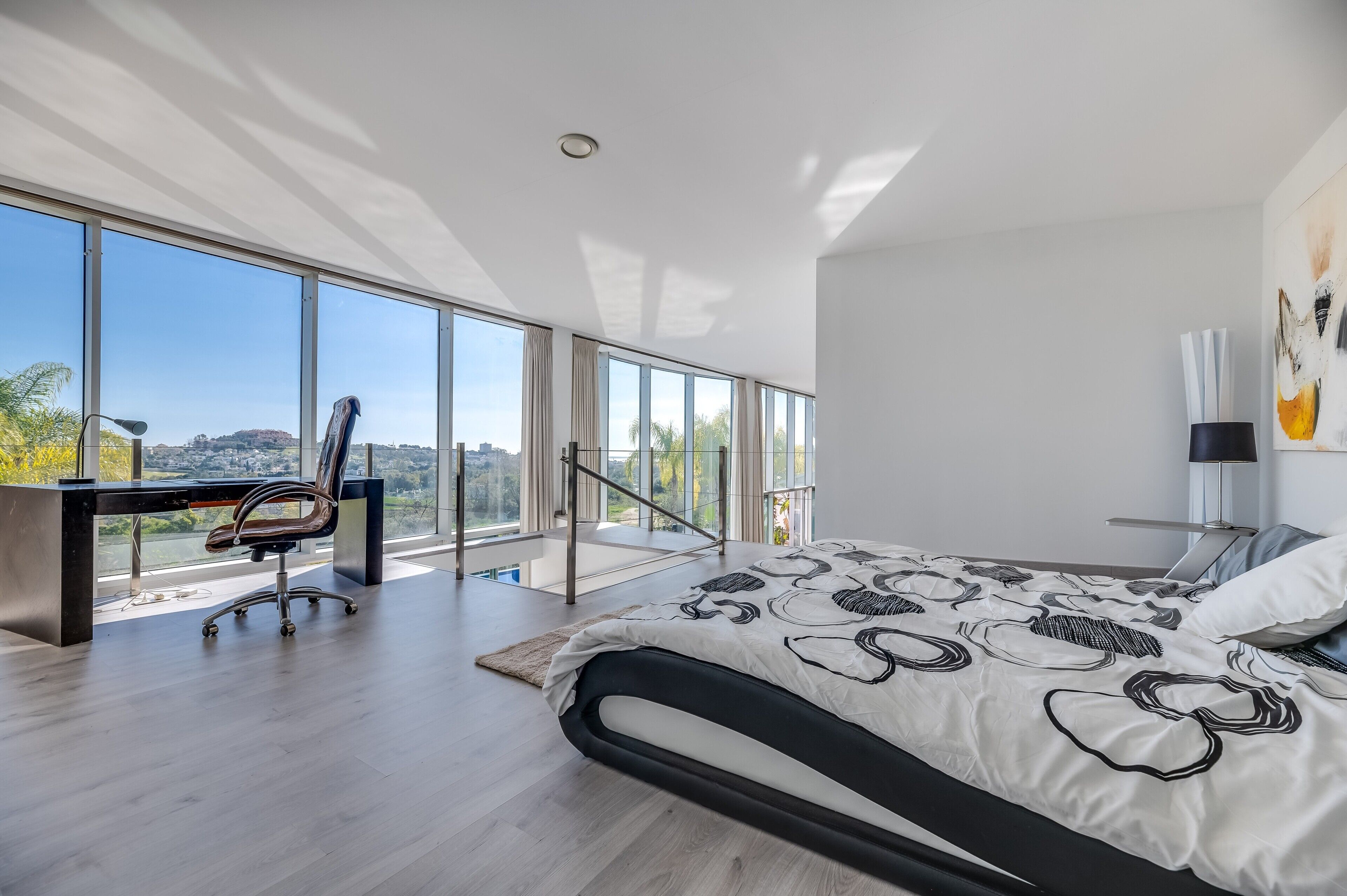 A modern bedroom with a large bed, a desk, and a chair, with a city view through large windows.