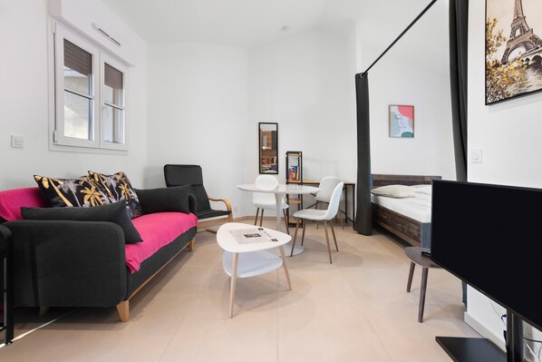 Ein modernes Wohnzimmer mit einem schwarzen Sofa, einem weißen Couchtisch und einem Fernseher. Es gibt einen Essbereich mit einem weißen Tisch und Stühlen sowie ein Bett mit einem schwarzen Kopfteil.