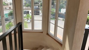 A corner room with three windows, a staircase, and a view of a gated area with greenery.