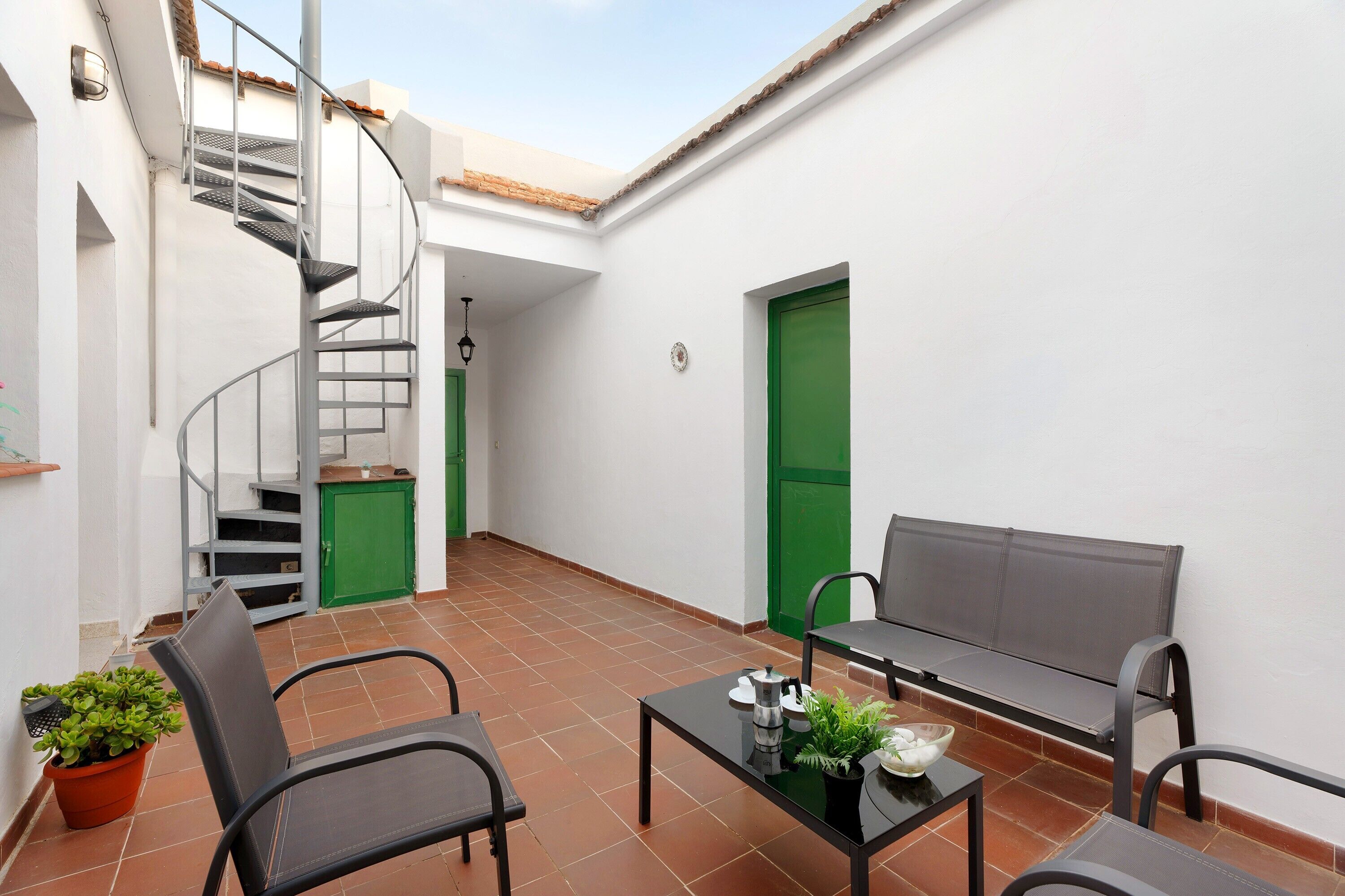 Une cour intérieure avec un escalier en colimaçon, une porte verte, un banc et une table agrémentée d’une plante.