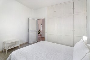 Un dormitorio con cama, un armario blanco, un estante pequeño blanco y una puerta que da a otra habitación.