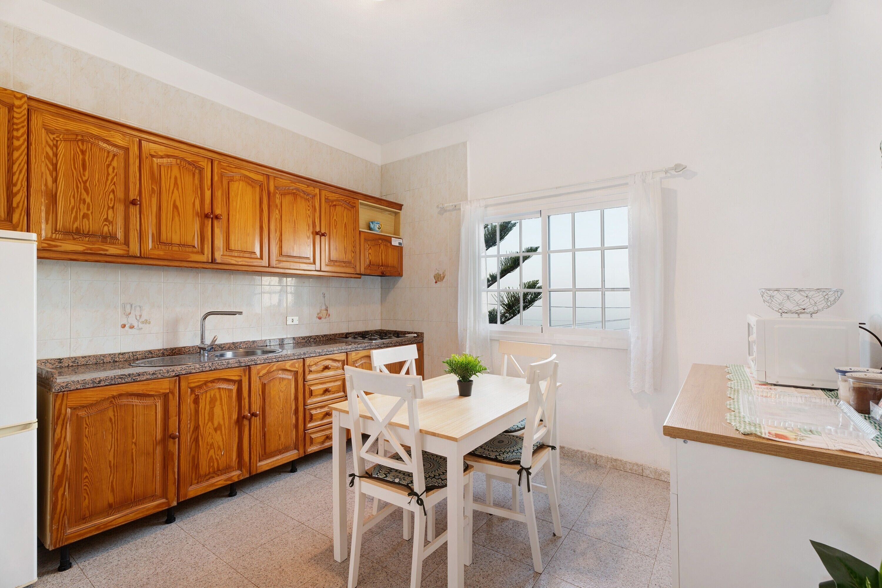 A kitchen with wooden cabinets, a granite countertop, a white dining table with chairs, and a window with curtains.