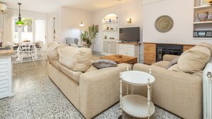 Ein Wohnzimmer mit Sofa, Couchtisch und Fernseher. Es gibt einen Kamin, einen Essbereich mit Stühlen und eine Küchenzeile mit Arbeitsplatte und Schränken.