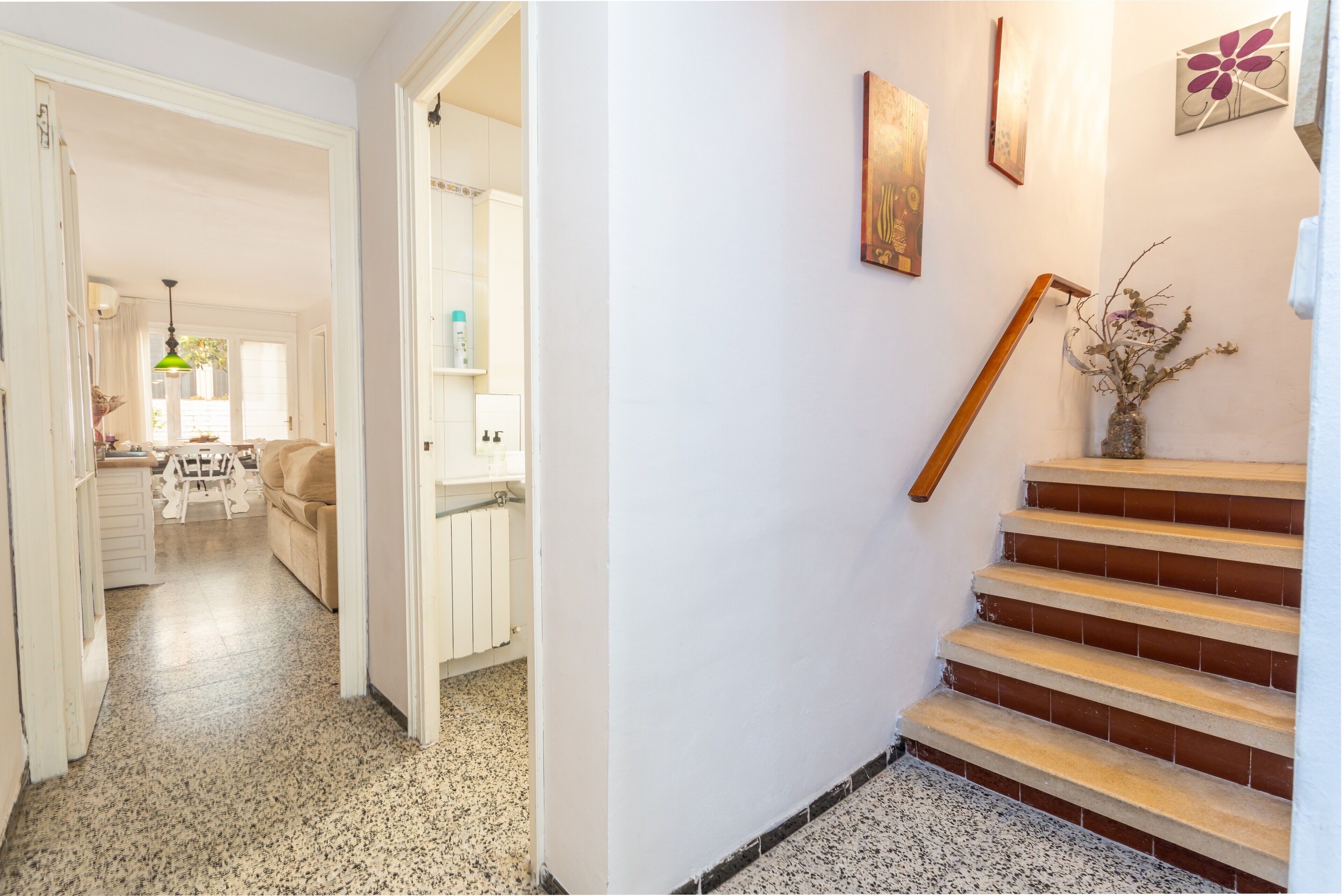 A hallway leading to a kitchen with a dining area, a staircase with a wooden handrail, and a vase with branches on the wall.
