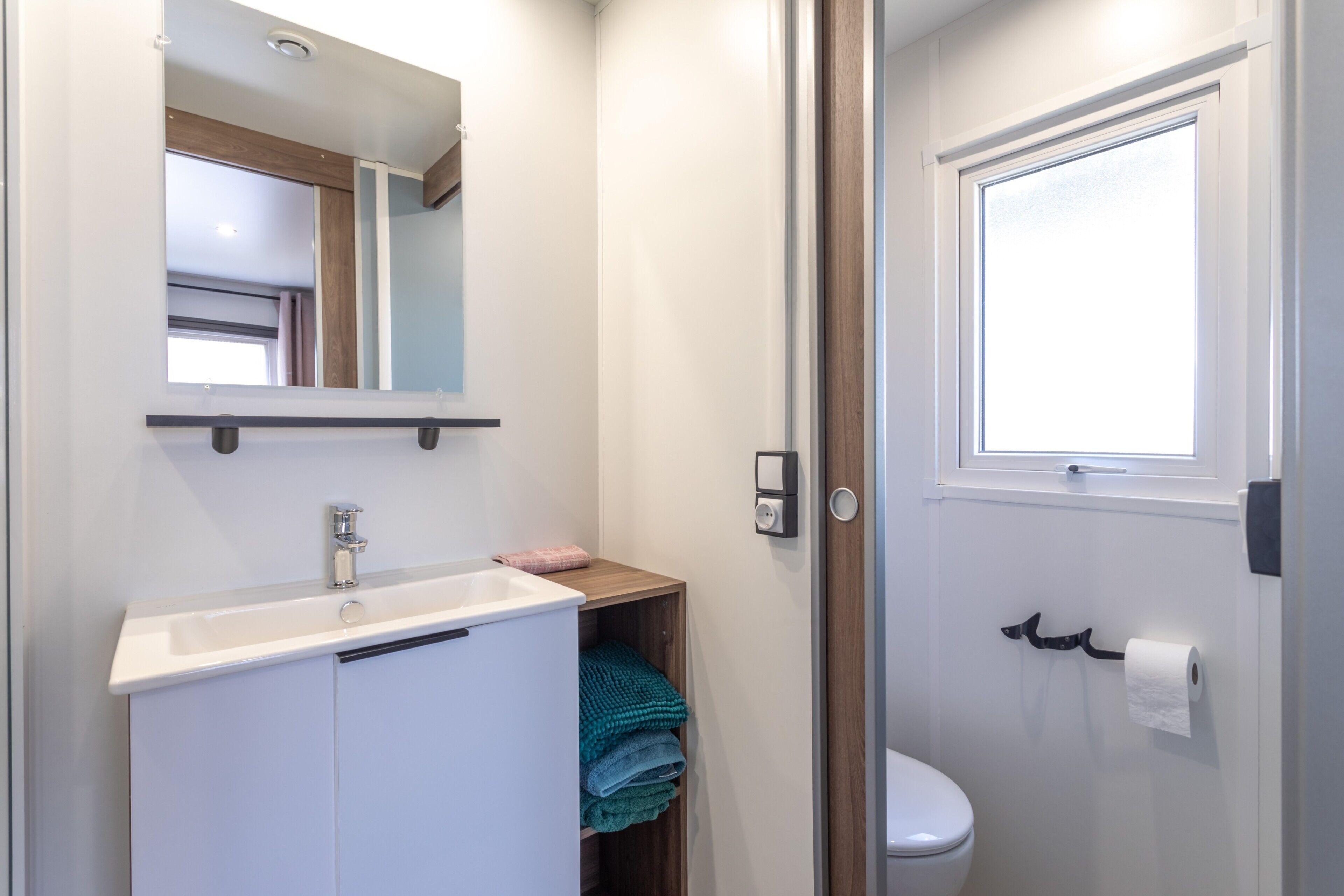 A bathroom with a white sink, mirror, and toilet. There is a window above the toilet and a towel rack with towels.