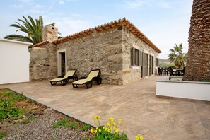 Un edificio de piedra con techo de tejas, un patio con sillones de descanso y una palmera.