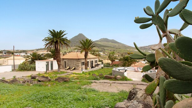 Un bâtiment coiffé d’un toit en tuiles, entouré de cactus et de palmiers, avec une montagne en arrière-plan.