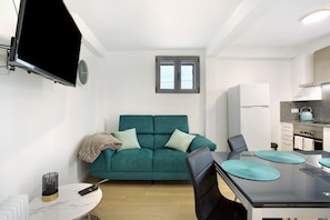 Un espacio habitable moderno con un sofá verde azulado, una mesa de comedor, un televisor montado en la pared y una zona de cocina.