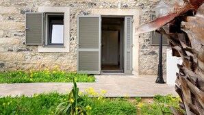 Un bâtiment en pierre avec deux portes ouvertes, une fenêtre avec des volets et un jardin où poussent des fleurs jaunes.