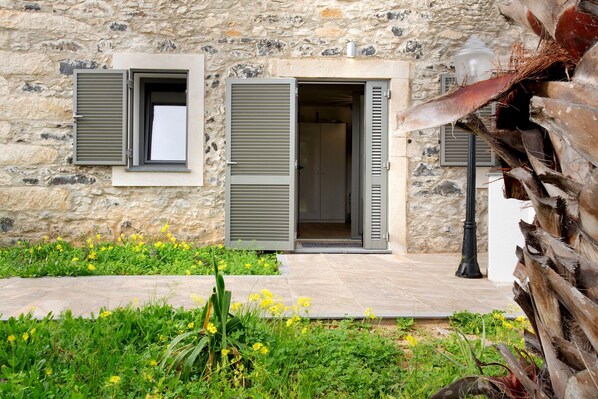 Un edificio de piedra con dos puertas abiertas, una ventana con contraventanas y un jardín con flores amarillas.