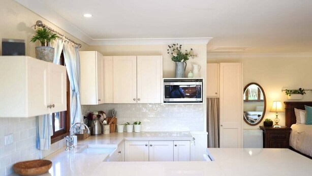 A modern kitchen with white cabinetry, a built-in microwave, and a sink. There is a dining area with a table and chairs, and a small plant on the table. - Fair Linden's Morrow (Bowral)