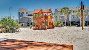 A beachside playground with a wooden play structure, a sandy beach, and a row of buildings in the background. - Port of Call~2BR~Sleeps 10~Dogs ok~Steps to beach (Port St. Joe)