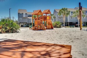 Een speeltuin aan het strand met een houten speelconstructie, een zandstrand en een rij gebouwen op de achtergrond.