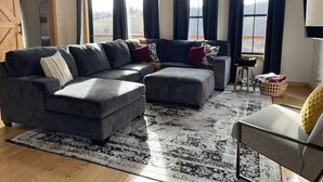 A modern living room with a sectional sofa, a small table, and a large window with curtains. - Bluebird Hill - rolling hills, meadows, large deck, fire pit, 3 bedroom, 2 bath (Guysville)