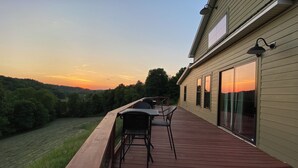 A deck with outdoor furniture, a view of a sunset, and a house with large windows. - Bluebird Hill - rolling hills, meadows, large deck, fire pit, 3 bedroom, 2 bath (Guysville)