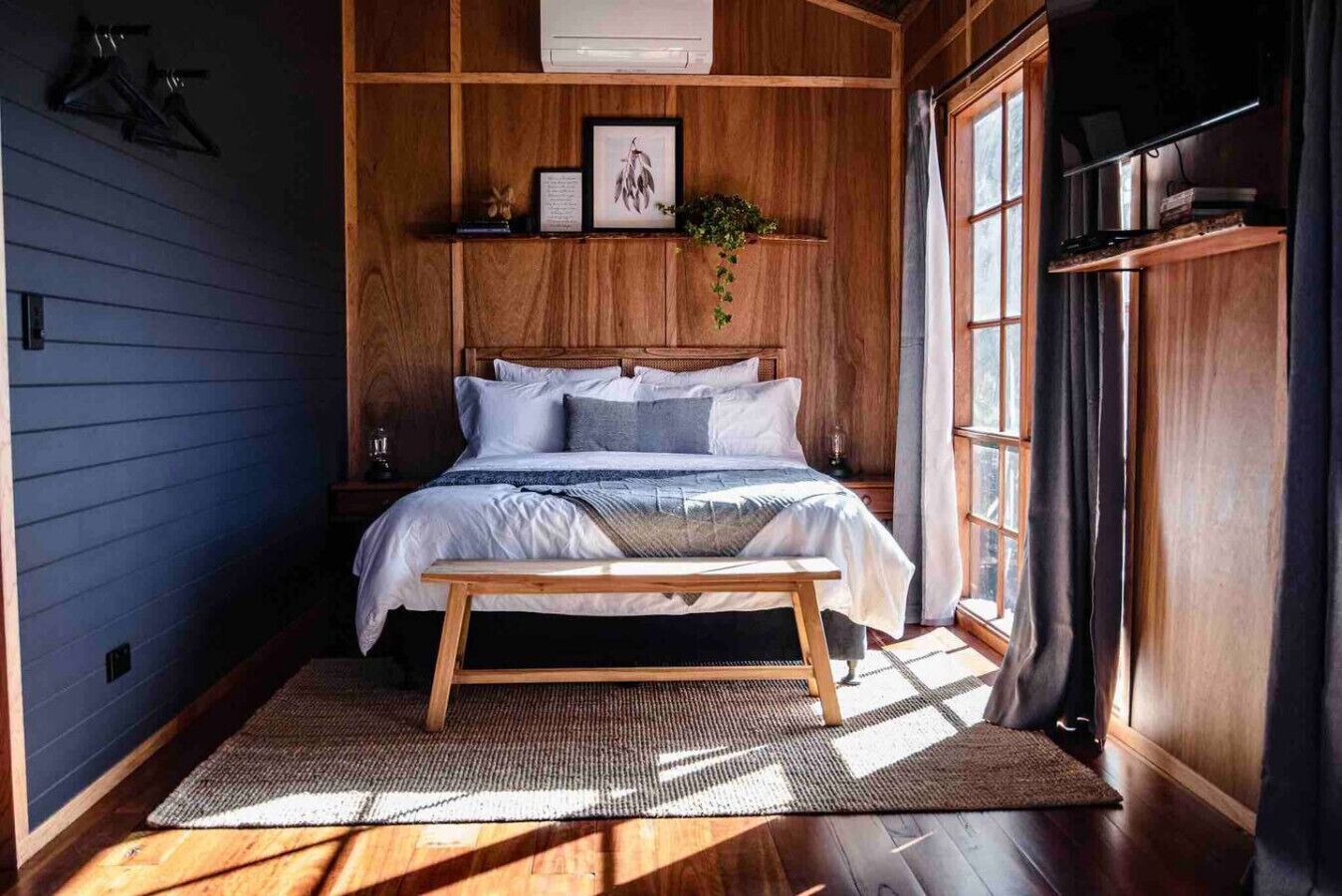 Un dormitorio con cabecera de madera, una cama con ropa de cama blanca, una mesita de noche y una ventana con cortinas.