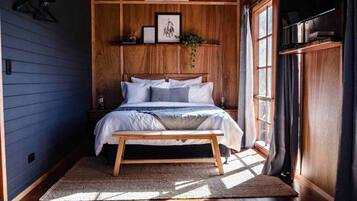 Un dormitorio con cabecera de madera, una cama con ropa de cama blanca, una mesita de noche y una ventana con cortinas.