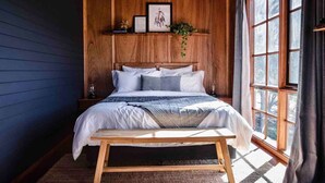 A bedroom with a wooden headboard, a bed with white and gray bedding, a bedside table with a lamp, and a window with curtains. - Grey Gum Luxury Cottage & Highland Cows (Mount Pleasant)