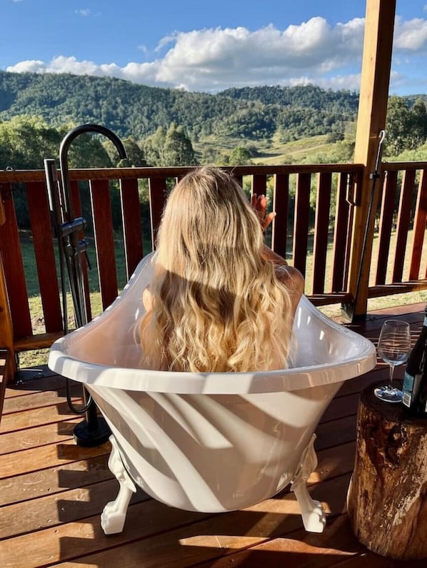 A person sitting in a white clawfoot bathtub on a wooden deck with a scenic mountain view in the background.
