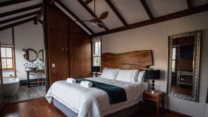 Un grand lit avec du linge de lit blanc et une couverture sombre, une tête de lit en bois, des tables de chevet avec des lampes, un grand miroir et une vue sur une salle de bain avec une baignoire et un lavabo.