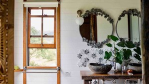 Une salle de bain avec une fenêtre à cadre en bois, un mur recouvert de carreaux blancs, un miroir orné d’un motif floral, un lavabo avec un receveur en pierre et une plante en pot.