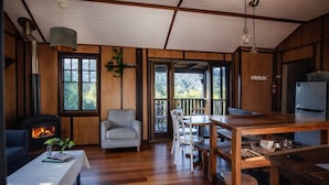 L’intérieur chaleureux d’un chalet, avec un poêle à bois, un coin repas et une grande fenêtre donnant sur le paysage extérieur.