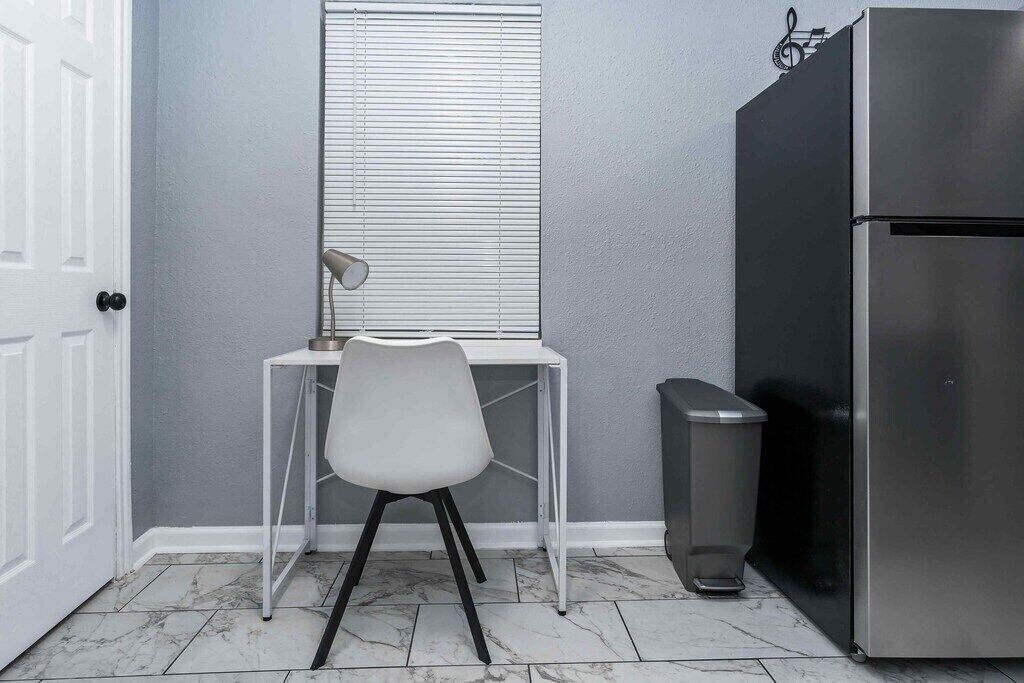 Een kleine keuken met een wit bureau, een stoel, een raam met zonwering, een roestvrijstalen koelkast en een afvalbak.