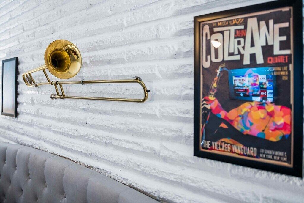 A brass trombone mounted on a white brick wall, a framed poster of Coltrane's 'A Love Supreme' album, and a tufted gray sofa.