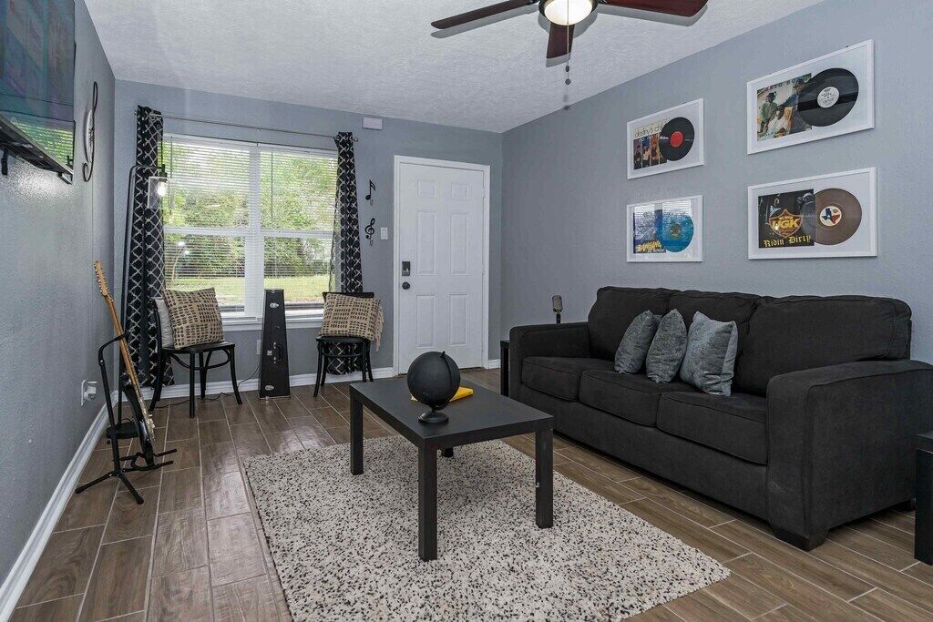 A living room with a dark grey sofa, a black coffee table, and a patterned rug. There are framed records on the wall and a guitar leaning against the wall.