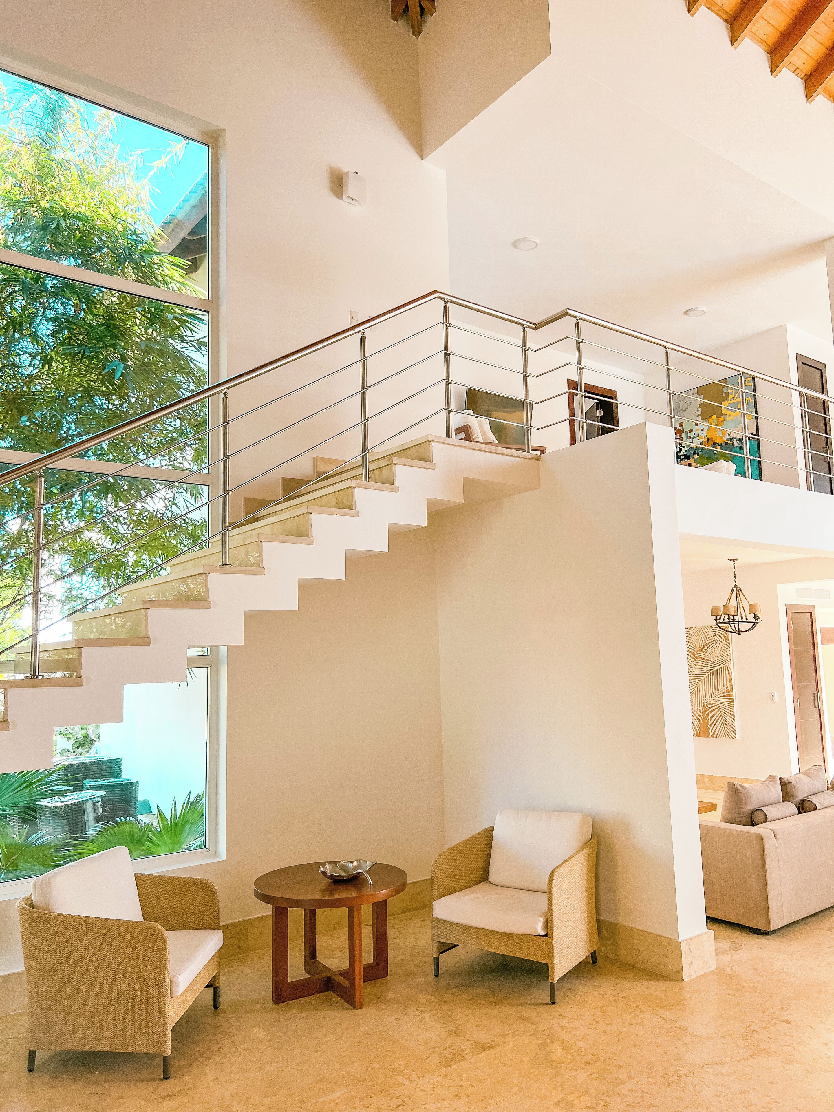 A spacious living area with a high ceiling, a staircase leading to an upper level, and large windows offering a view of greenery.