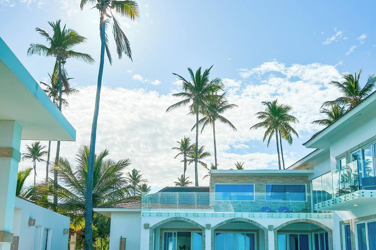 A pool area with lounge chairs and a dining area, surrounded by palm trees and a two-story building.
