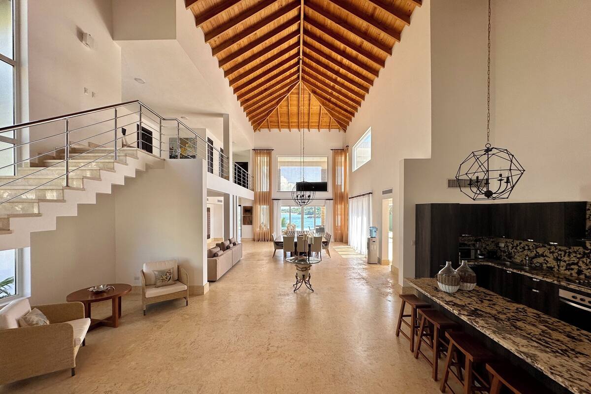 A spacious interior with a high wooden ceiling, a central seating area, a kitchen with bar stools, and a staircase leading to an upper level.